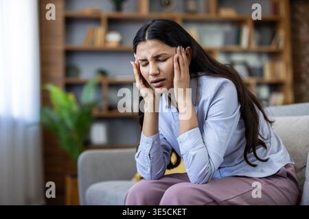 Junge Frau, die mit geschlossenen Augen auf einem Sofa sitzt, schmerzhafte Kopfschmerzen oder Migräne erlitten hat, Hände an die Schläfen drückt und Unwohlsein, Müdigkeit und Stress zeigt Stockfoto