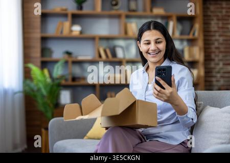 Junge Frau, die auf dem Sofa sitzt und während eines Videoanrufs mit ihrem Smartphone eine Lieferung auspackt, lächelt und einen Neukauf in entspannter Atmosphäre zeigt Stockfoto