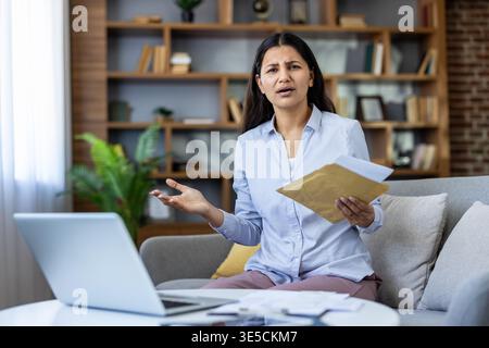 Frau, die auf einem Sofa sitzt und sich besorgt und gestresst fühlt, während sie einen Umschlag mit Rechnungen und Finanzdokumenten öffnet und frustriert über die unerwarteten schlechten Nachrichten in Bezug auf Finanzen schaut Stockfoto