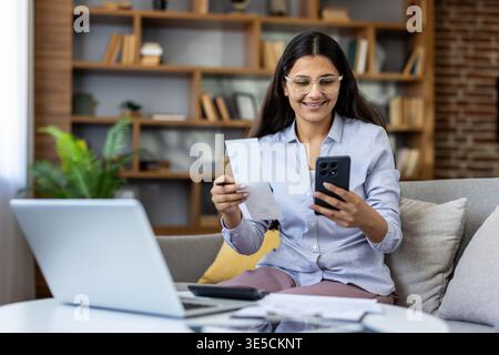 Junge indianerin überprüft Belege auf dem Smartphone, führt Berechnungen für Steuern oder Budgetplanung durch, verwendet moderne digitale Banking- und Buchhaltungswerkzeuge zu Hause Stockfoto
