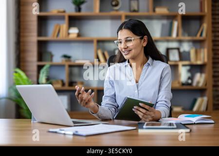 Frau mit Brille, die einen Videoanruf auf ihrem Laptop führt, ein Notebook hält und Gesten bewegt, während sie an ihrem Schreibtisch in einem Heimbüro sitzt, sich an Fernarbeit und Online-Kommunikation beteiligt Stockfoto