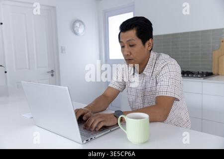 Asiatischer Mann, der an der Küchentheke sitzt und auf silbernem Laptop mit blassgrünem Becher in der Nähe tippt Stockfoto