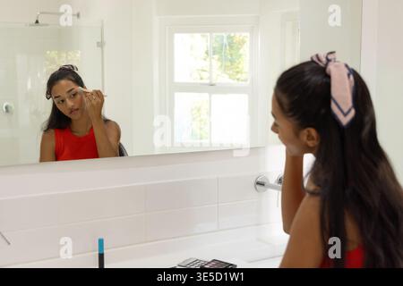 Asiatische Frau, die Make-up am Waschbecken im Bad aufträgt, Pinsel hält, Spiegel überprüft, rotes Oberteil trägt. Kosmetik, Reflexion, Lippenstift, Kosmetikbereich, Tageslicht, waschen Stockfoto