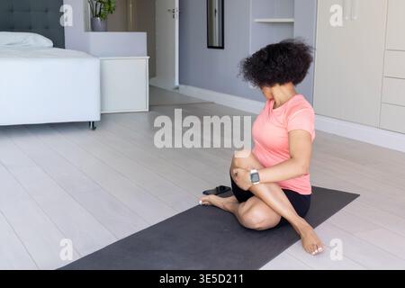 Midadult afroamerikanische Dame, die auf Yogamatte im Schlafzimmer rosa Oberteil mit Smartwatch, Kopierraum dreht. Frau, Stretch, Balance, Wellness, Zuhause, ruhig, Stockfoto