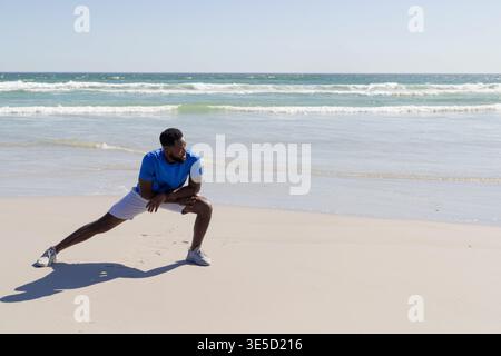 Mittelgroßer afroamerikanischer Mann, der seitlichen Ausfallschritt am Strand mit blauem Hemd und weißen Schuhen macht. Athlet, Stretch, Outdoor, Küste, Küste, oc Stockfoto