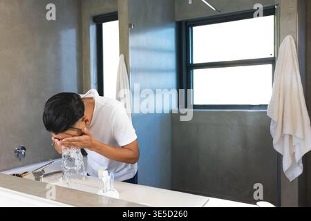 Mann beugt sich über Waschbecken, schröpft Wasser und spritzt Gesicht, läuft Wasserhahn und aktiviert Gesundheitsgrafiken. Badezimmer, Waschanlage, Fenster, Routine, Hygiene, Kosmetikbereich, f Stockfoto