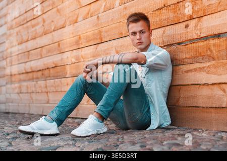 Ein stilvoller, hübscher Studentenmann mit einer Frisur in einer Denim-Kleidung mit Hemd, Jeans und Sneakers sitzt auf dem Boden nahe einer Holzwand Stockfoto