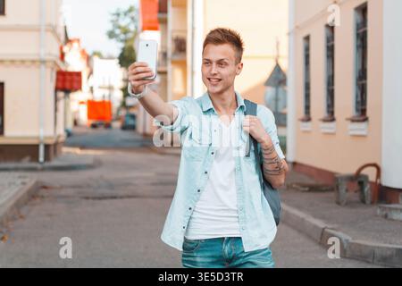 Ein glücklicher, trendiger Student, hübscher Kerl mit einem Hairstyle in modischer, lässiger Denim-Kleidung und Rucksack macht ein Selfie-Foto auf einem Smartphone und geht Stockfoto