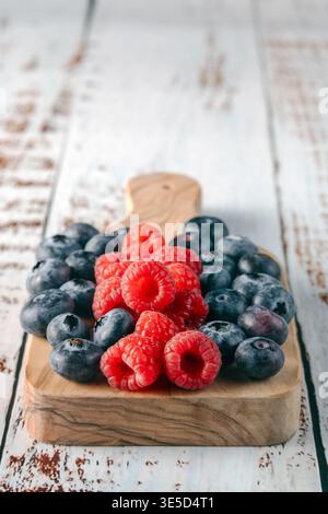 Blick von oben auf frische Himbeeren und Heidelbeeren auf einem kleinen hölzernen Schneidebrett über einem weißen rustikalen Hintergrund Stockfoto