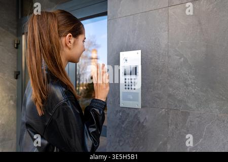 Eine Junge Frau Benutzt Die Wohnung-Gegensprechanlage Draußen. Eine junge Frau in einer schwarzen Lederjacke benutzt eine Außensprechanlage an einem modernen Wohneingangseingang für Stockfoto