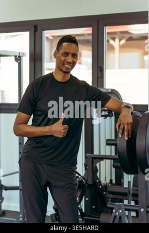afroamerikanischer Mann, der sich in einem modernen Fitnessstudio auf Gewichte stützt, Sportbekleidung trägt, selbstbewusst lächelt und eine Daumen-nach-oben-Geste macht, die achi symbolisiert Stockfoto