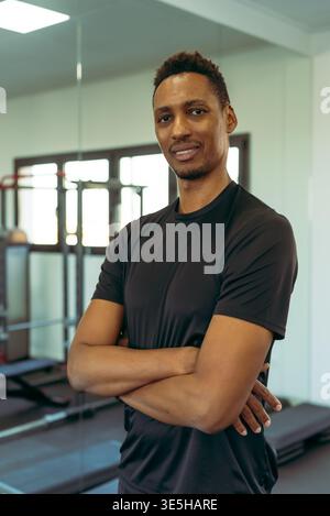Junger athletischer schwarzer Mann, der mit gekreuzten Armen steht und Selbstvertrauen und eine positive Einstellung in einem modernen Fitnessraum ausstrahlt und den persönlichen Zug repräsentiert Stockfoto
