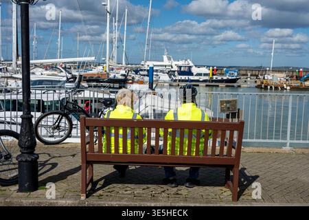 Ein älteres Paar, das gemeinsam auf einer Bank am Yachthafen im Yarmouth Harbour an der Küste der isle of wight, Großbritannien, unterwegs ist Stockfoto