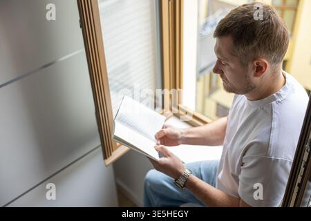 Blick auf den Mann, der Buch für Fenster liest, mit sanftem Tageslicht und neutralem Innenraum Stockfoto