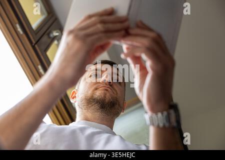 Niedriger Blickwinkel eines Mannes, der ein Buch hält und in der Nähe des Fensters liest, bei weichem Tageslicht Stockfoto