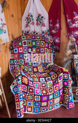 Farbenfroher handgefertigter afghanischer Holzsessel mit Häkelbezug neben floralen, gestickten Textilien und antikem Spinnrad in gemütlichem traditionellem Haus setti Stockfoto