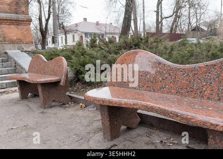 Aufwendig geschnitzte Bänke aus rotem Granit mit welligen Formen stehen in einem öffentlichen Park, umgeben von Sträuchern, Ziegelbauten und blattlosen Bäumen und Stockfoto