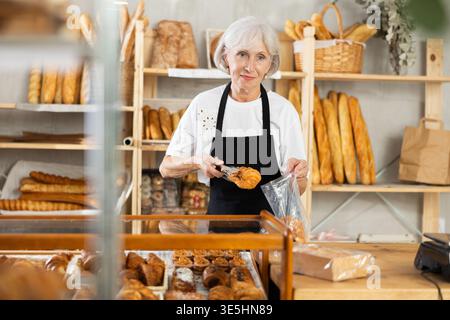 Die Senior-Verkäuferin bringt Croissant in eine Plastiktüte für den Käufer, verpackt Waren. Stockfoto