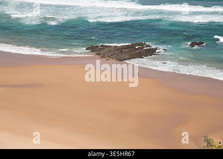 Breiter Sandstrand Mit Türkisfarbenen Wellen Und Felsigem Felsvorsprung, Lone Walker On Shore Stockfoto