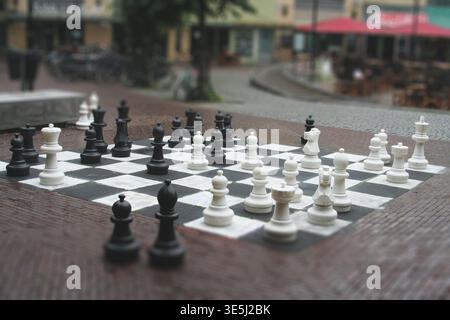 Ein großes Schachbrett mit schwarz-weißen Kunststoffteilen auf einem Kopfsteinpflaster in einer öffentlichen Fußgängerzone mit verschwommenem Hintergrund Stockfoto