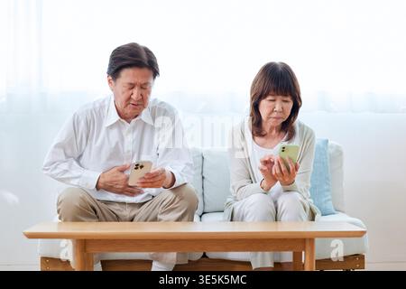 Japanisches Seniorpaar besorgt über Gesundheit Mann mit Magenschmerzen mit Smartphones auf Sofa Stockfoto