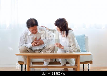 Ein japanischer Seniorenmann leidet unter Brustschmerzen und seine Frau tröstet ihn auf dem Wohnzimmer-Sofa Stockfoto