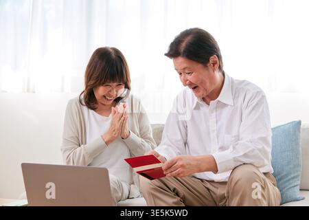 Glückliches japanisches Seniorenpaar, das zu Hause ein Passbuch und einen Laptop sucht, um Geld zu sparen Stockfoto