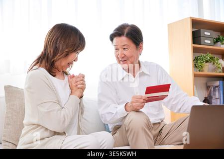 Lächelndes japanisches Seniorpaar, das über zukünftige Finanzpläne mit Bankbuch und Laptop zu Hause diskutiert Stockfoto
