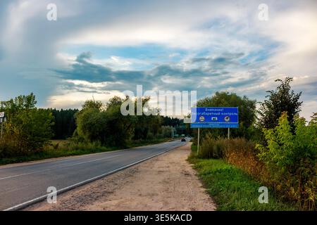 Ein russisches Verkehrsschild warnt die Fahrer vor einem bevorstehenden Kontrollpunkt für Gewicht und Übergröße, umgeben von üppig grünen Sommerlaub unter einem dramatisch bewölkten Himmel. Stockfoto