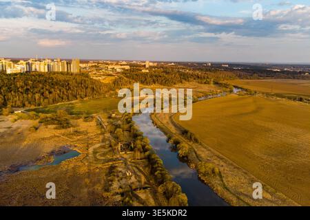 Atemberaubender Blick aus der Luft, der einen sich windenden Fluss mit einem Abendhimmel einfängt, umgeben von goldenen Feldern und der entfernten Skyline der Stadt im Frühlingslicht Stockfoto