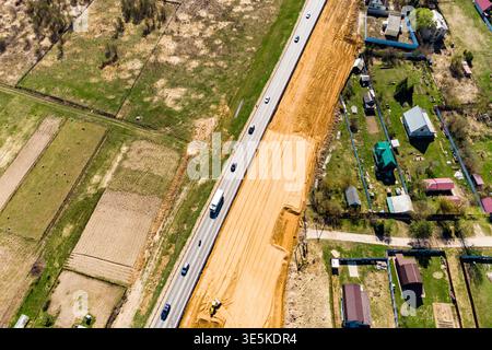 Die Luftaufnahme erfasst ein Autobahnausbauprojekt neben ländlichen Häusern und grünen Feldern im Frühjahrsentwicklungsgebiet Stockfoto