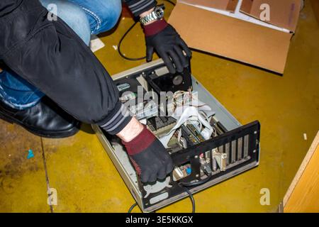 Handschuhbekleidete Hände sind damit beschäftigt, einen alten Desktop-PC in einer düsteren, industriell wirkenden Umgebung zu entfernen. Alte Computerteile werden gerettet oder auf einem Gelb gesaugt Stockfoto