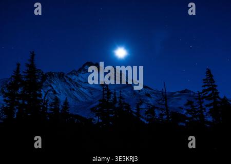 Eine Bergkette hat eine Silhouette vor einem dunklen Himmel mit hellem Mond. Die Szene ist ruhig und friedlich, der Mond strahlt einen sanften Glanz auf den Schnee Stockfoto