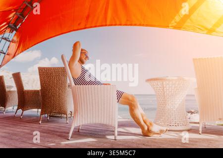 Mann Entspannung Strand entspannter Mann sitzt in einem Korbsessel auf einer Terrasse mit Blick auf das Meer an einem sonnigen Tag Stockfoto