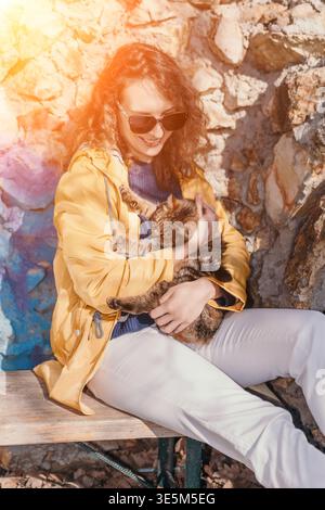 Frau Katze, die junge Frau in gelber Jacke umarmt, kuschelt eine Tabbykatze draußen an einem sonnigen Tag, die an einer Steinmauer sitzt Stockfoto