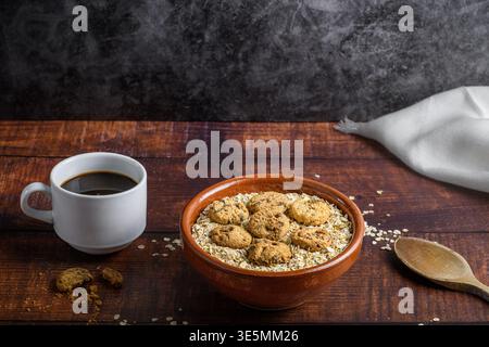 Rustikale Frühstücksszene mit schwarzem Kaffee, Haferflocken und Schokoladenkeksen in einer Tonschale, Holzlöffel und Leinenserviette auf dunklem Holz. Gemütliche hausgemachte M Stockfoto