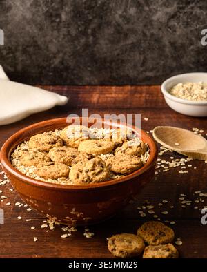 Seitenansicht einer rustikalen Schüssel gefüllt mit Haferflocken und Schokoladenkeksen auf einem dunklen Holztisch, mit Löffel, Leinentuch und extra Hafer. Gemütlich hausgemacht Stockfoto