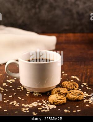 Vertikales rustikales Frühstücksbild mit schwarzem Kaffee und Schokoladenkeksen auf dunklem Holz, umgeben von Haferflocken und weichen Leinenstoff. Minimal hausgemacht Stockfoto