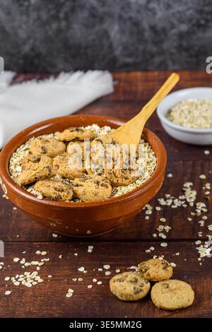 Vertikale Frühstücksszene mit rustikaler Tonschale mit Haferflocken und Schokoladenkeksen, Holzlöffel und extra Keksen im Vordergrund auf dunklem Holz. Stockfoto