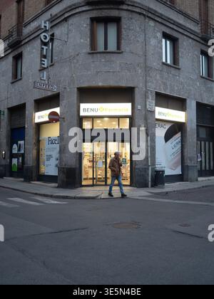 Lombardei, Italien - 15. März 2026 Ein einsamer Mann überquert die Straße vor einer BENU Farmacia und einem Hoteleingang auf einer europäischen Stadtstraße Stockfoto