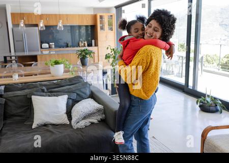 Lächelnde afroamerikanische Mutter und Tochter, die sich in einem modernen Wohnzimmer umarmen und Topfpflanzen zeigen Stockfoto