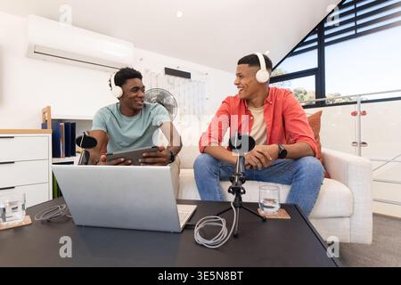 Aufnahme verschiedener männlicher Freunde, die im Heimstudio auf der Couch sitzen, Kopfhörer tragen und mit einem Laptop arbeiten Stockfoto