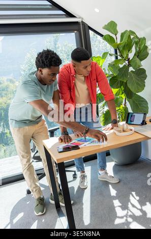 Verschiedene männliche Kollegen, die sich über den Schreibtisch beugen und Farbpalettenmuster und Smartphone im Studio untersuchen Stockfoto