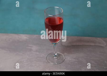 Ein klares Glas enthält ein hellrotes Getränk, das auf einer hellen Oberfläche platziert ist. Der Hintergrund ist blau und die Szenerie scheint drinnen zu sein. Licht fällt auf das Getränk. Stockfoto