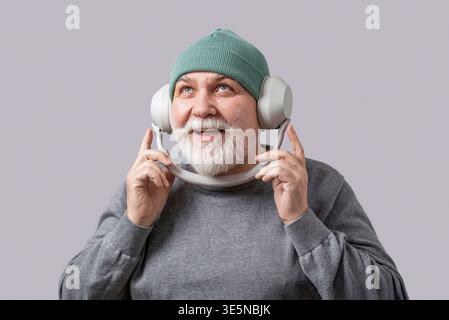 Älterer Rentner hört Musik, tanzt, trägt Kopfhörer, isoliert auf grauem Hintergrund. Porträt eines lächelnden reifen bärtigen Mannes, der zuhört Stockfoto