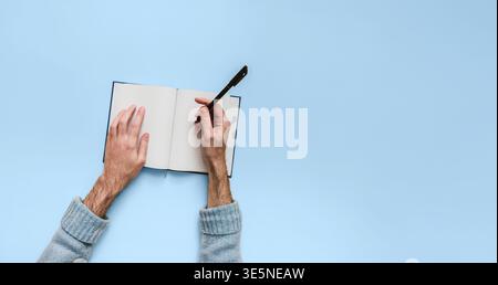 Männliche Hände schreiben mit schwarzem Stift in offenem, liniertem Notizbuch von oben auf einheitlichem blauem Hintergrund. Linke Hand hält Seite, großer weißer Bereich für Text Out Stockfoto