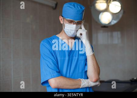 Arzt Chirurg in blauer Uniform und Brille steht und denkt im OP Stockfoto
