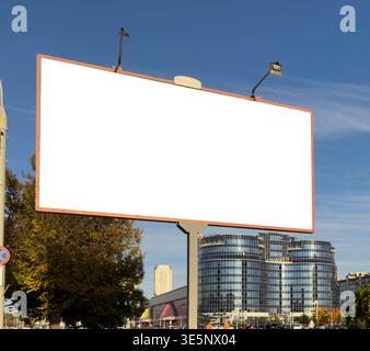 Große leere Plakatwand am Straßenrand vor blauem Himmel mit modernen Stadtgebäuden im Hintergrund. Leere weiße Werbefläche bietet eine klare Kopie Stockfoto