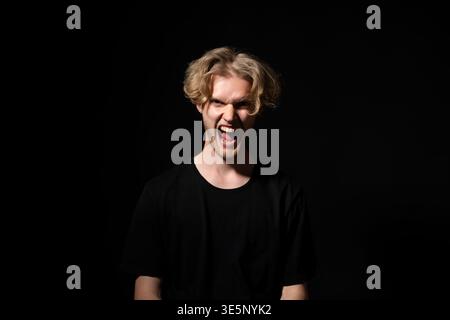 Junger Mann, der extremen Wut und Wut ausdrückt, mit weit geöffnetem Mund auf dunklem Hintergrund schreiend, Frustration, Stress und aggressive Emotionen zeigt Stockfoto