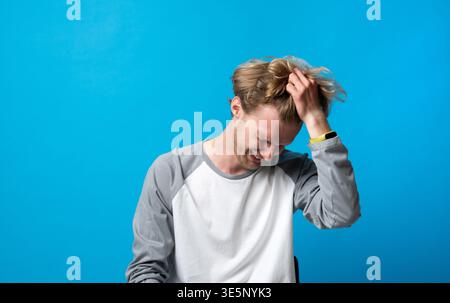 Junger Mann lacht und kratzt sich den Kopf aus Frustration oder Peinlichkeit, fühlt sich gestresst, verwirrt oder macht einen Fehler auf blauem Hintergrund Stockfoto